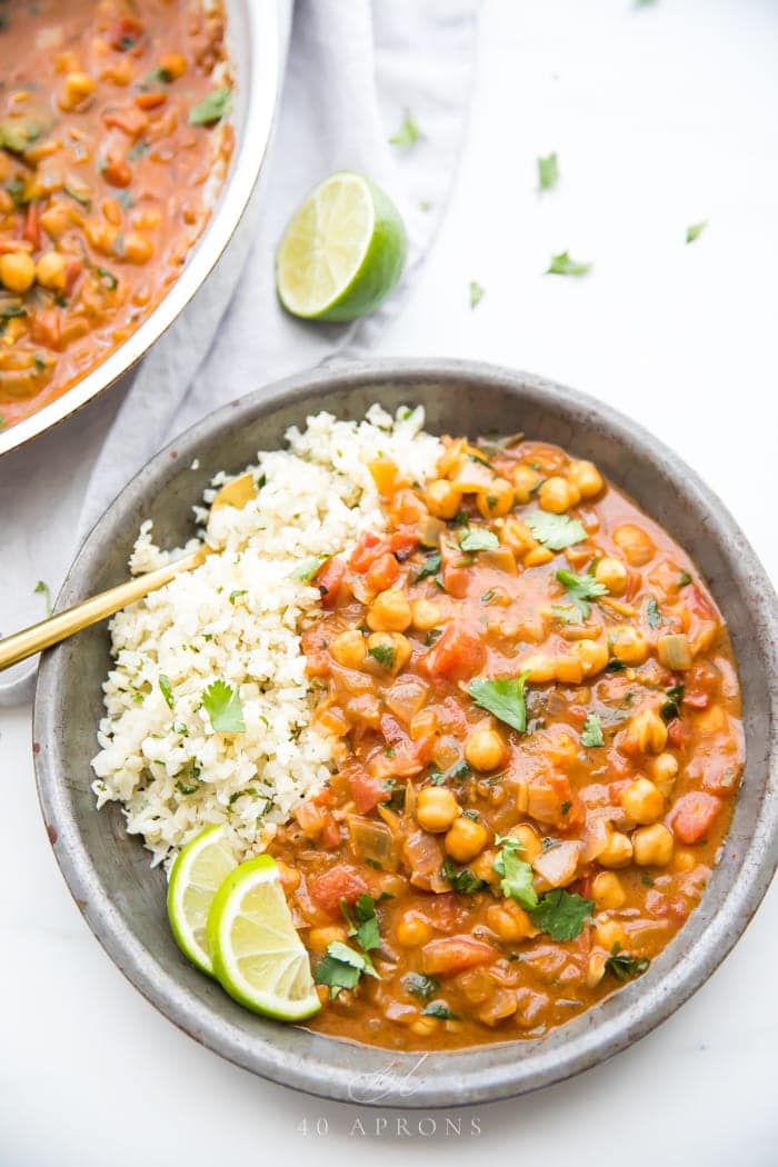 Overhead shot of creamy vegan coconut curry in a bowl with cauliflower rice, cilantro and lime wedge garnishes, and a skillet next to the bowl