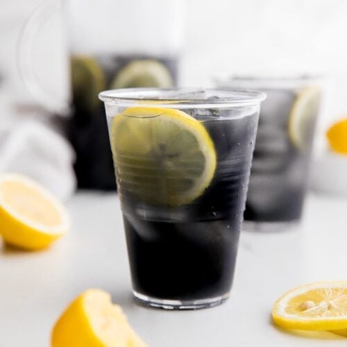 A glass of black charcoal lemonade with lemon slice, with glass and pitcher in background