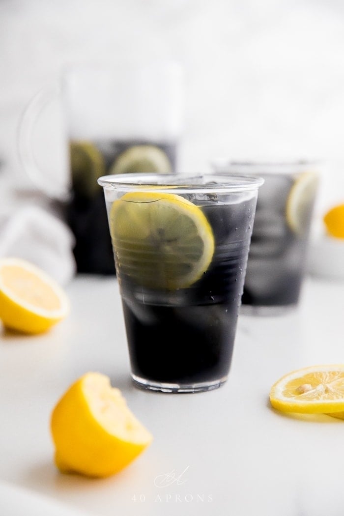 A glass of black charcoal lemonade with lemon slice, with glass and pitcher in background