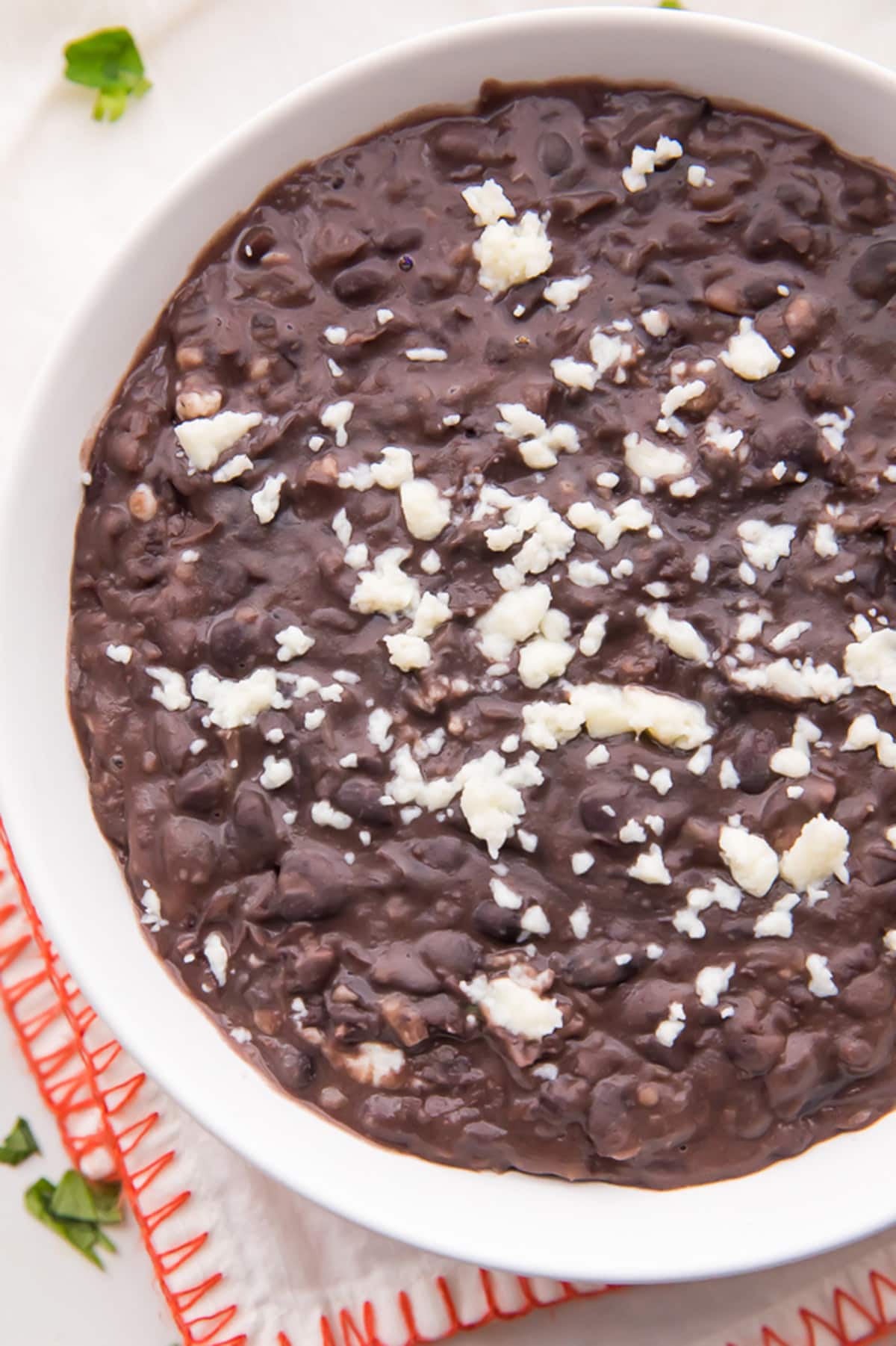 Refried black beans topped with cheese crumbles in a large white bowl resting on a red-edged clothed napkin.