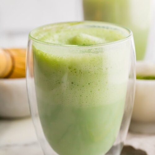 two matcha lattes in glass cups with a bamboo whisk to the side