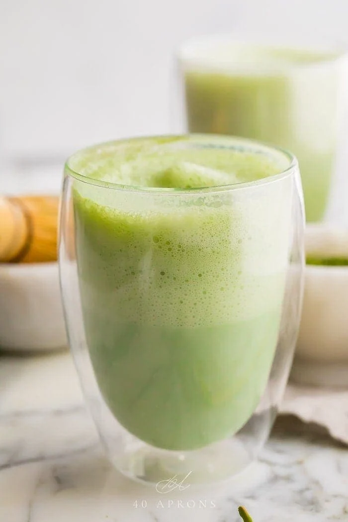 two matcha lattes in glass cups with a bamboo whisk to the side