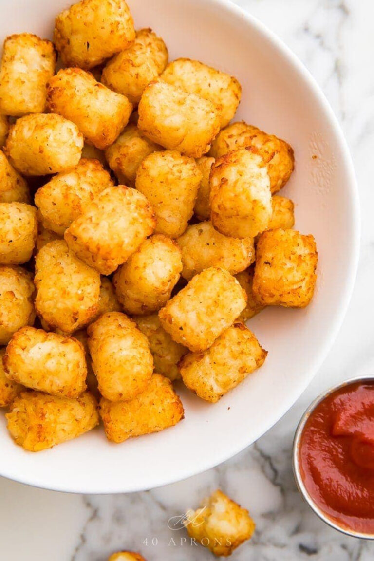 Crispy, golden air fryer tater tots in a large white serving bowl next to a small ramekin of bright red ketchup.