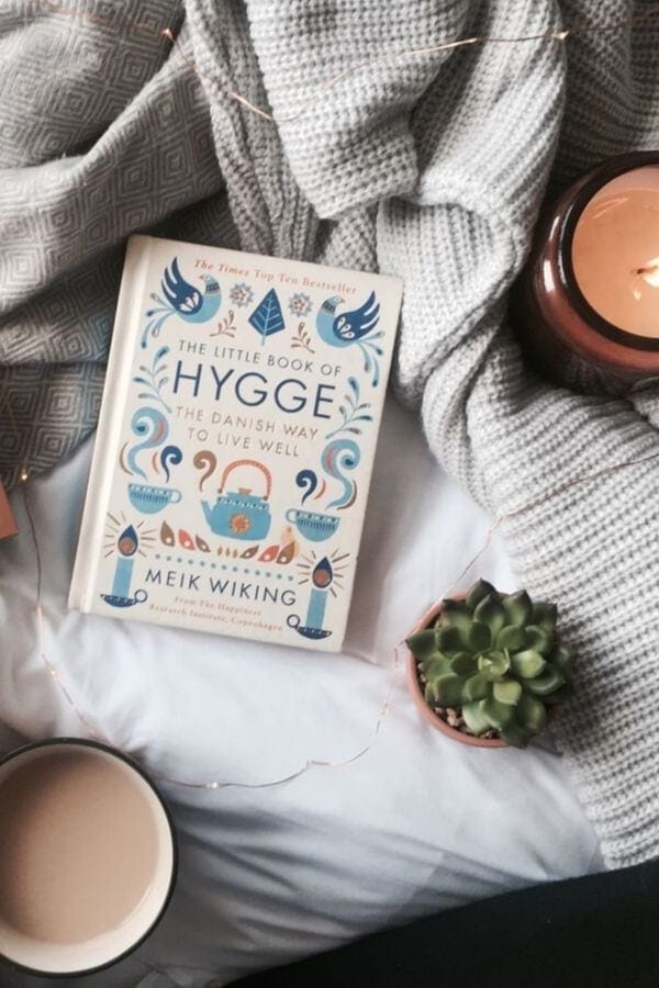 overhead shot of The Little Book of Hygge by Meik Wiking lying on a bed with a lit scented candle and a mug of coffee on the side