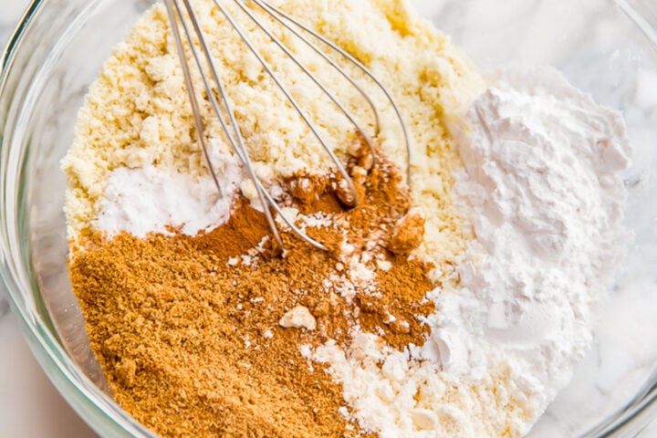 Dry ingredients for pumpkin muffins in a large glass mixing bowl with a silver whisk resting in the bowl.