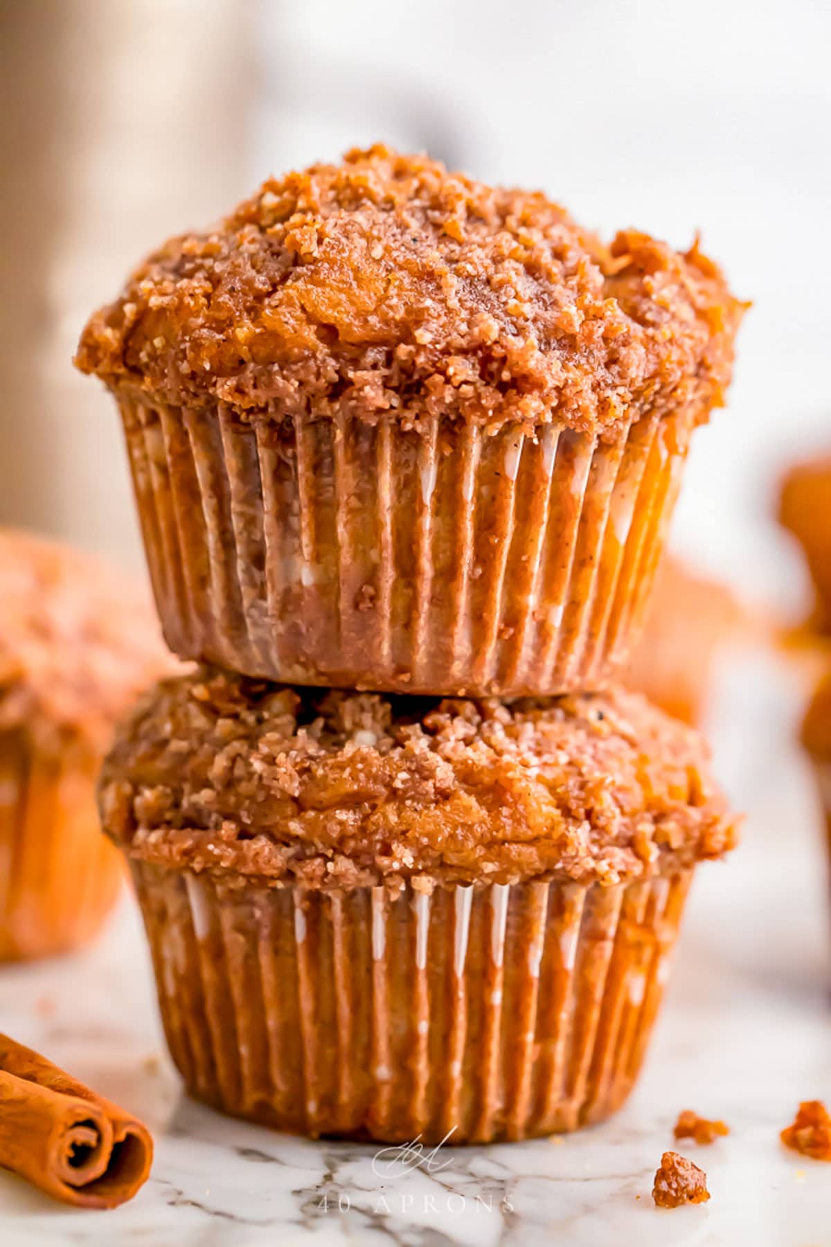 Two pumpkin muffins with a spiced crumb topping stacked on top of each other, with other muffins out of focus in the background.