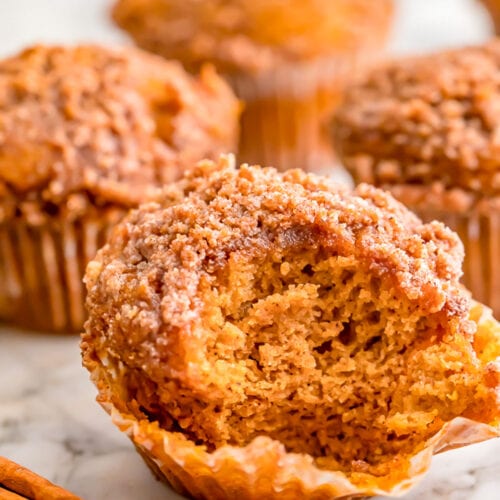 A pumpkin muffin, with the paper lining peeled down and a large bite missing from the front side of the muffin. More whole muffins stand in the background.