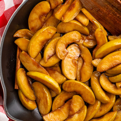 A large cast iron skillet of fried apple slices coated in brown sugar cinnamon butter resting on a table with a red and white checkerboard napkin.