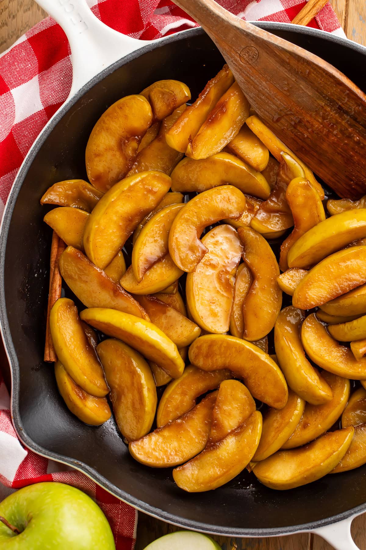 A large cast iron skillet of fried apple slices coated in brown sugar cinnamon butter resting on a table with a red and white checkerboard napkin.