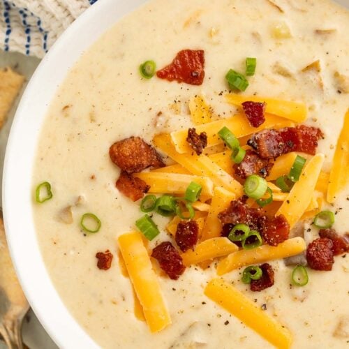 A large white bowl of creamy Crockpot potato soup topped with pieces of bacon and large shreds of cheddar cheese.