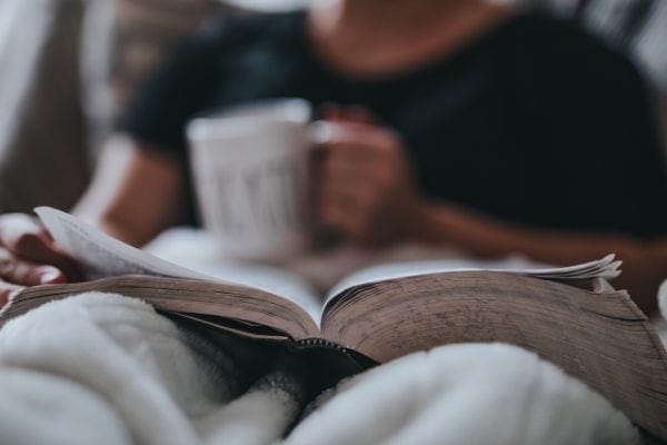 a man reading a book in bed with a mug of tea in one hand