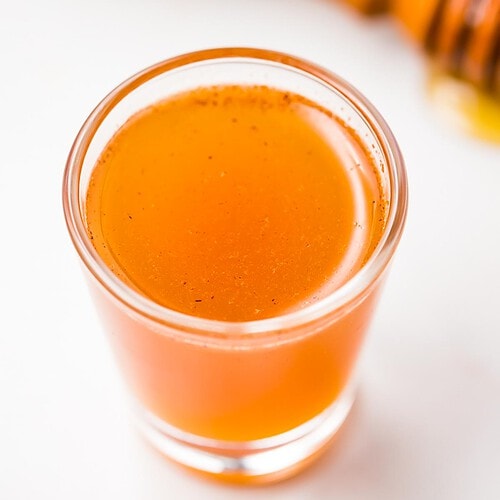 A clear glass shotglass holding an orange liquid peppered with cinnamon, in front of a honey wand dripping with honey.