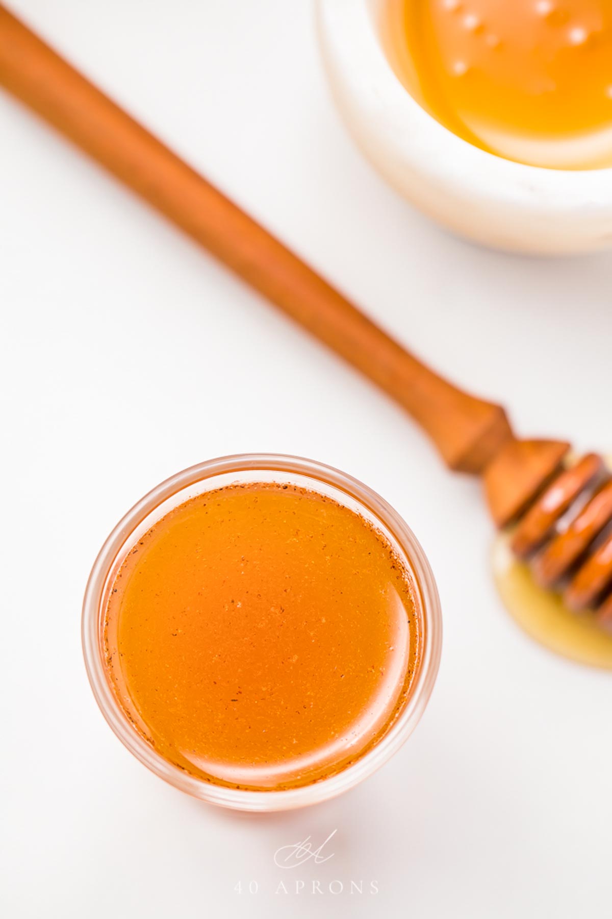 A clear glass shotglass holding an orange liquid peppered with cinnamon, in front of a honey wand dripping with honey.