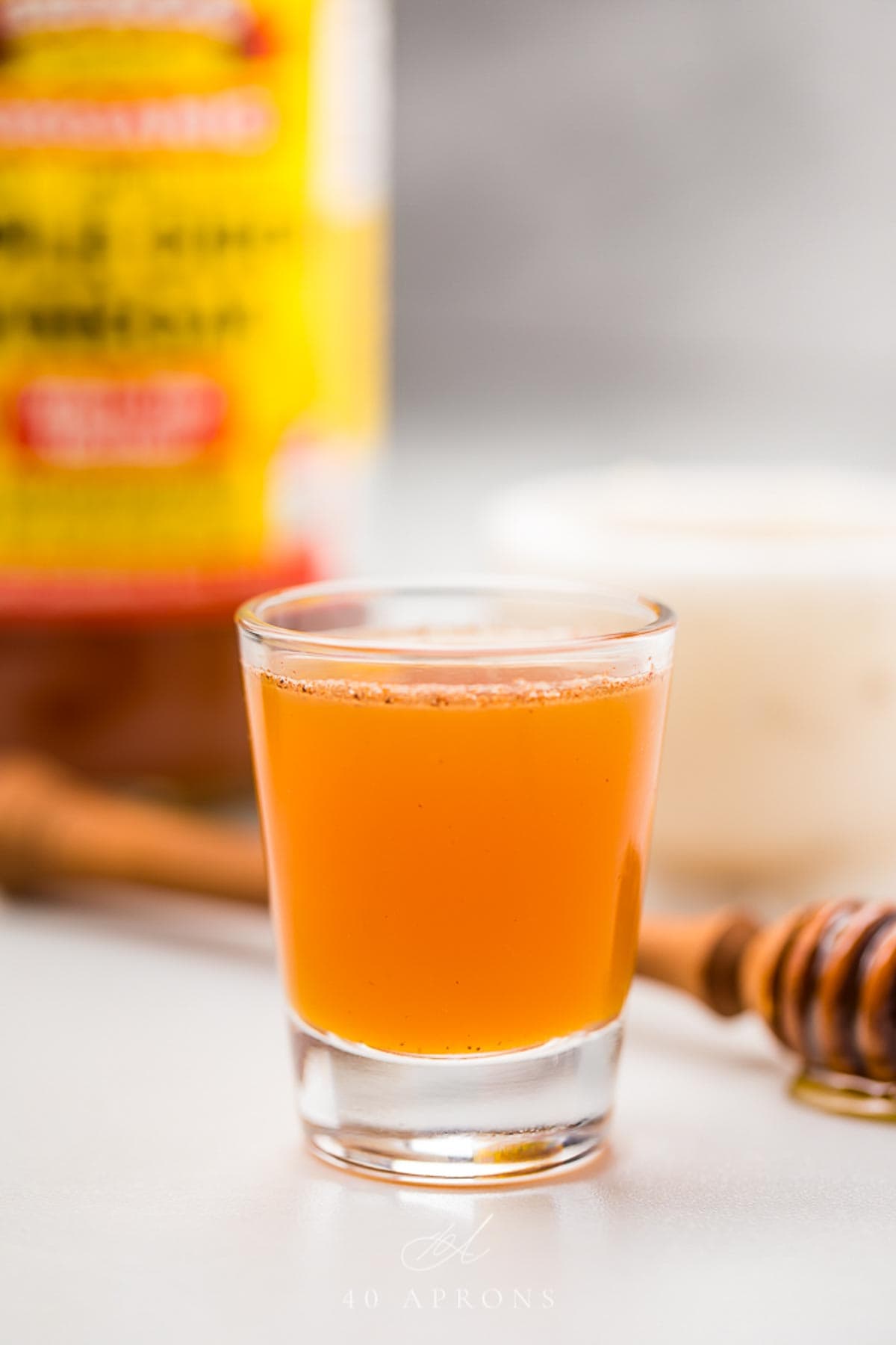 A clear glass shotglass holding an orange liquid peppered with cinnamon, in front of a honey wand dripping with honey.