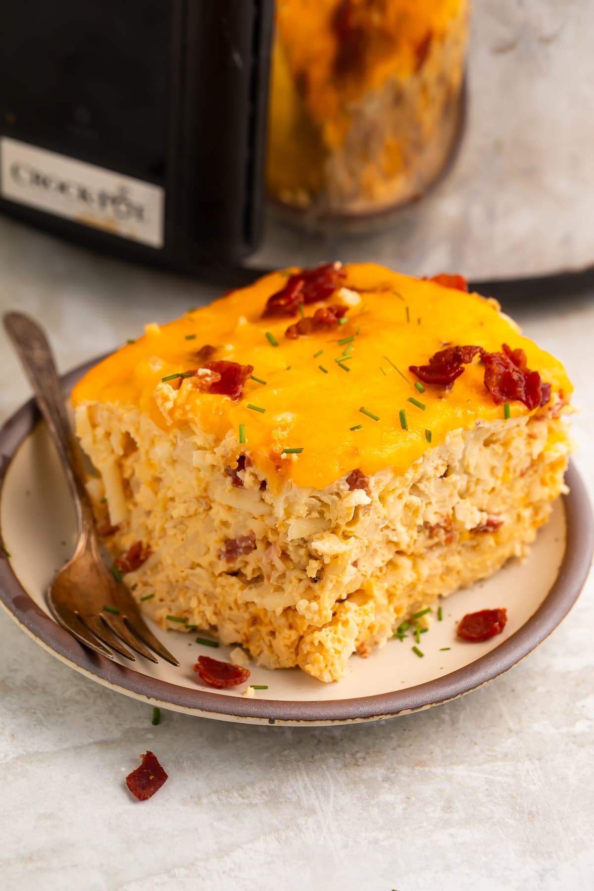 A square, multi-layered slice of Crockpot breakfast casserole on a plate in front of a slow cooker.