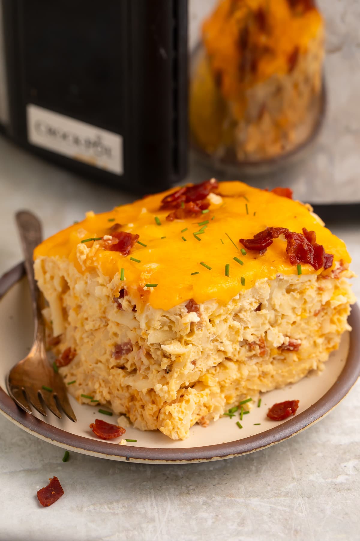 A square, multi-layered slice of Crockpot breakfast casserole on a plate in front of a slow cooker.