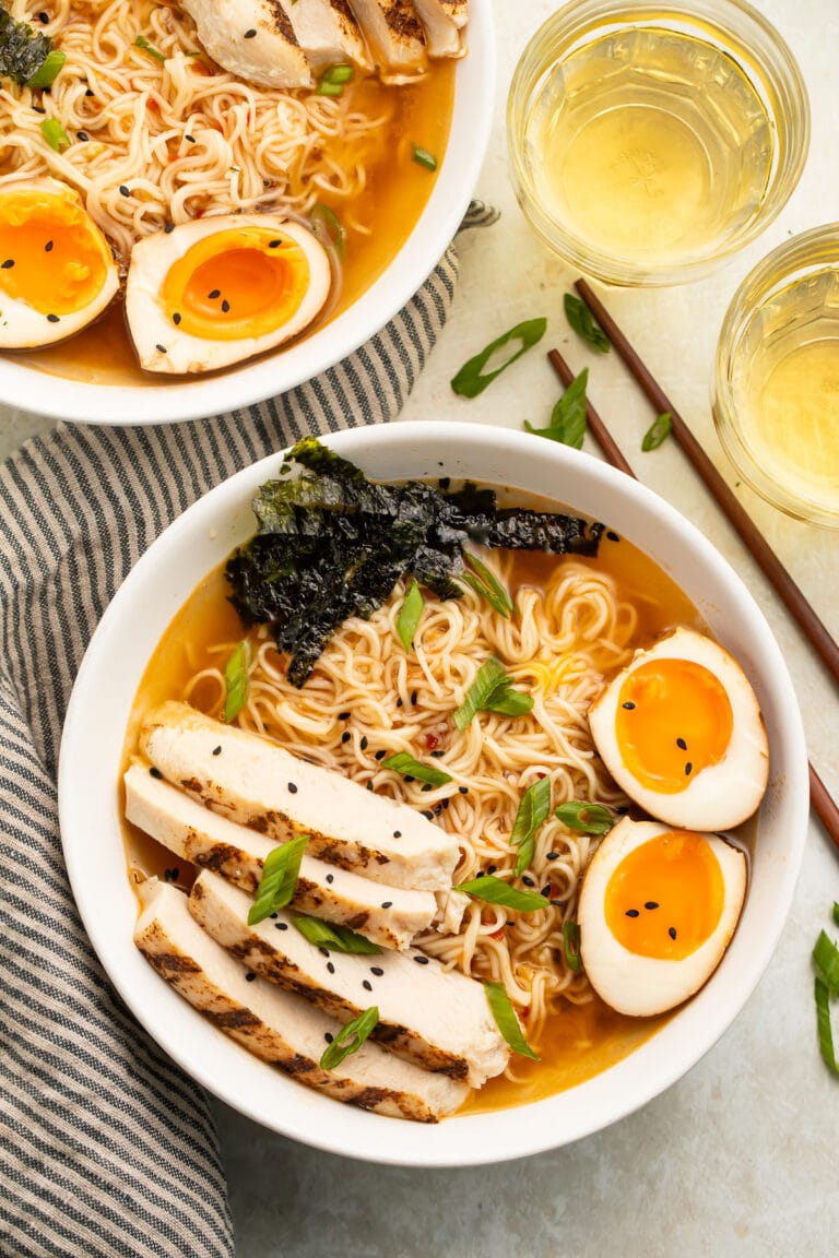 Top-down view of two white bowls holding spicy ramen noodles in broth with slices of chicken, two yolky egg halves, and green garnish.