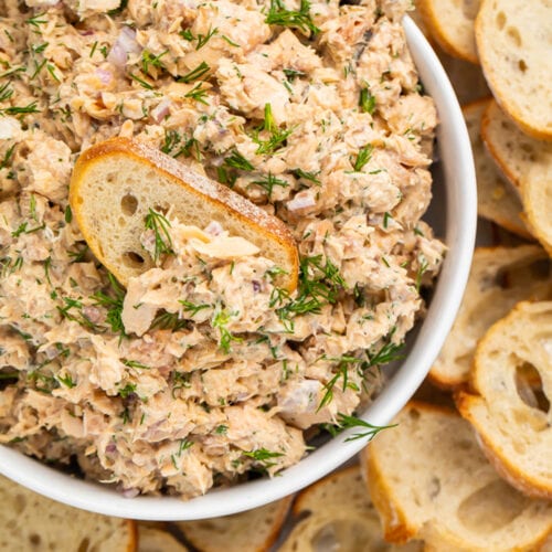 A bowl of canned salmon salad surrounded by crostini, with a slice of crostini in the dip.