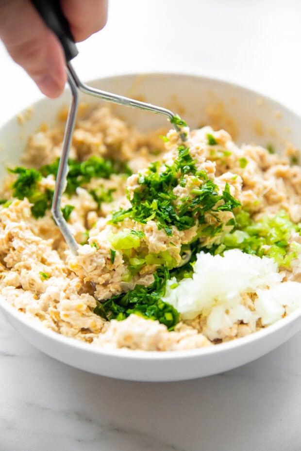 Chickpea salad ingredients in a large bowl with a potato masher