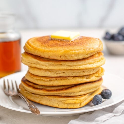 A stack of 6 paleo pancakes resting on a plate next to 2 blueberries. A pat of butter sits atop the pancakes.