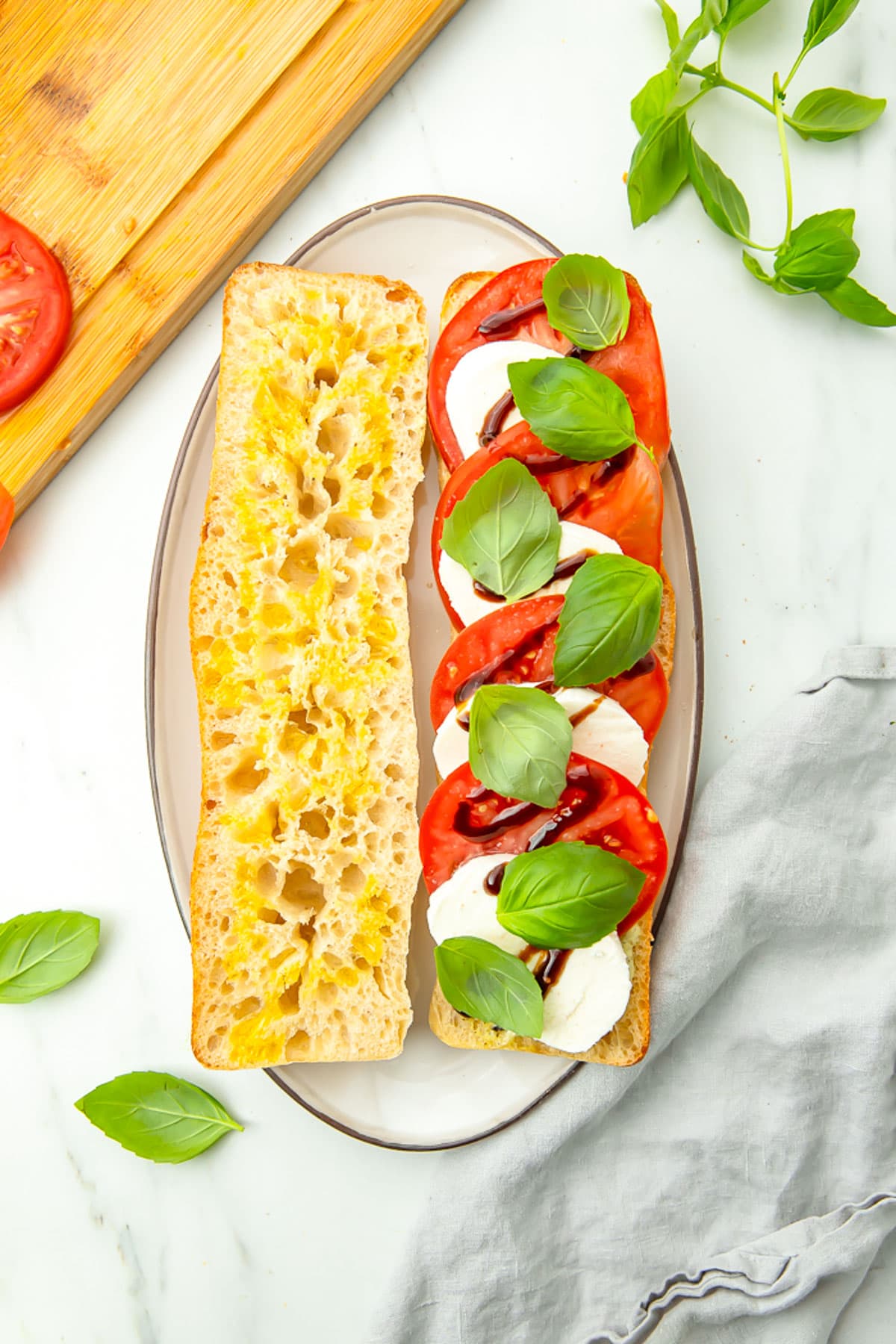 Top-down view of a caprese sandwich resting on a plate. The top piece of bread sits to the left of the bottom piece, showing alternating slices of mozzarella and tomato topped with basil and balsamic.