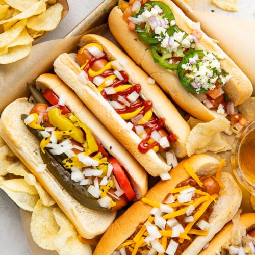 5 air fryer hot dogs in hot dog buns with various toppings arranged on a baking sheet.