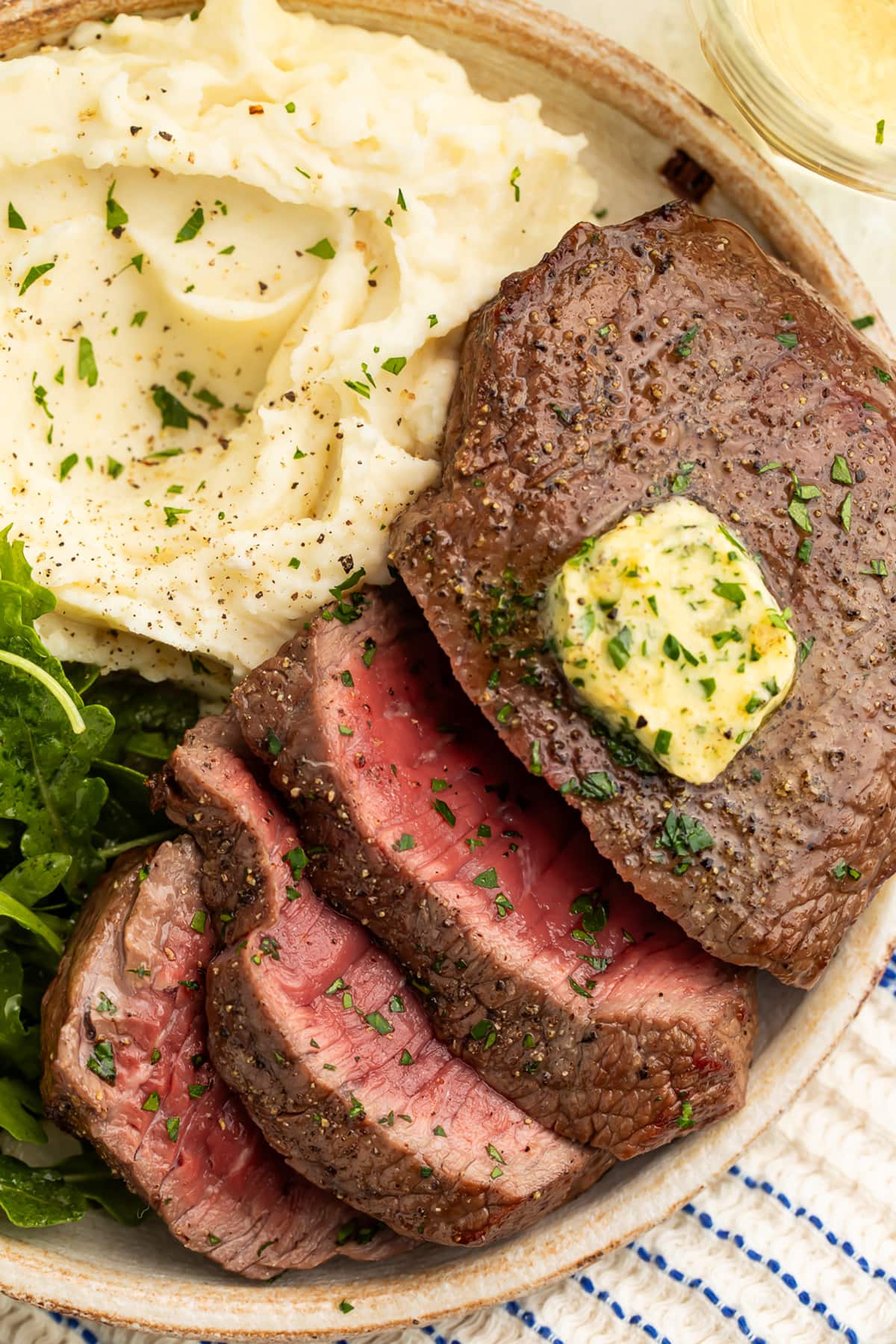 Close-up of medium-rare air fryer steak, topped with homemade herb butter, sliced and arranged on a plate with mashed potatoes and a small salad.