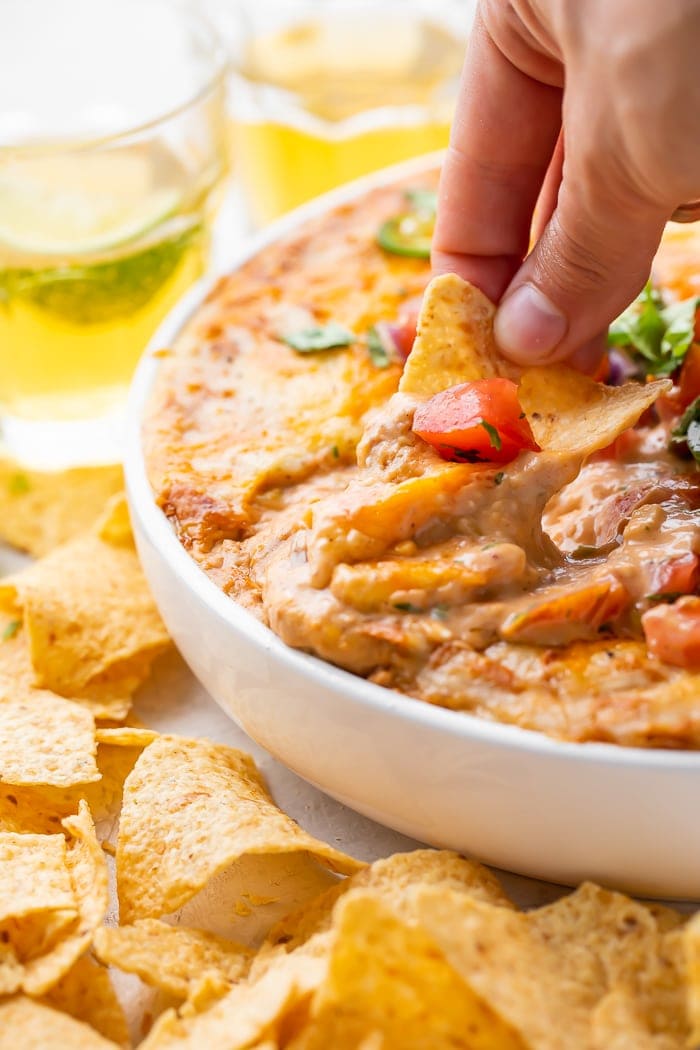 A hand dipping a corn chip into a bowl of bean dip in a bowl surrounded by glasses of beer and more chips