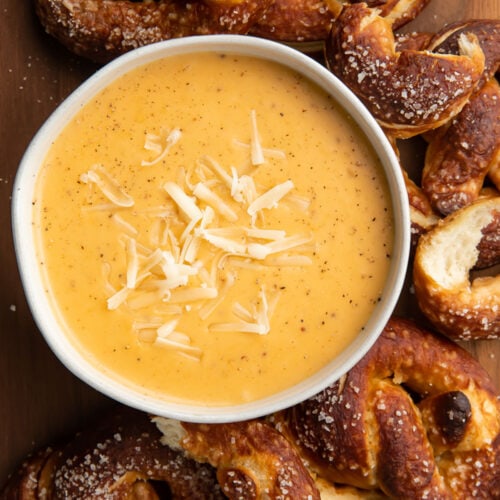 Overhead, top down look at a white bowl of rich yellow-orange beer cheese dip topped with shredded mozzarella surrounded by deep brown soft pretzels with salt.