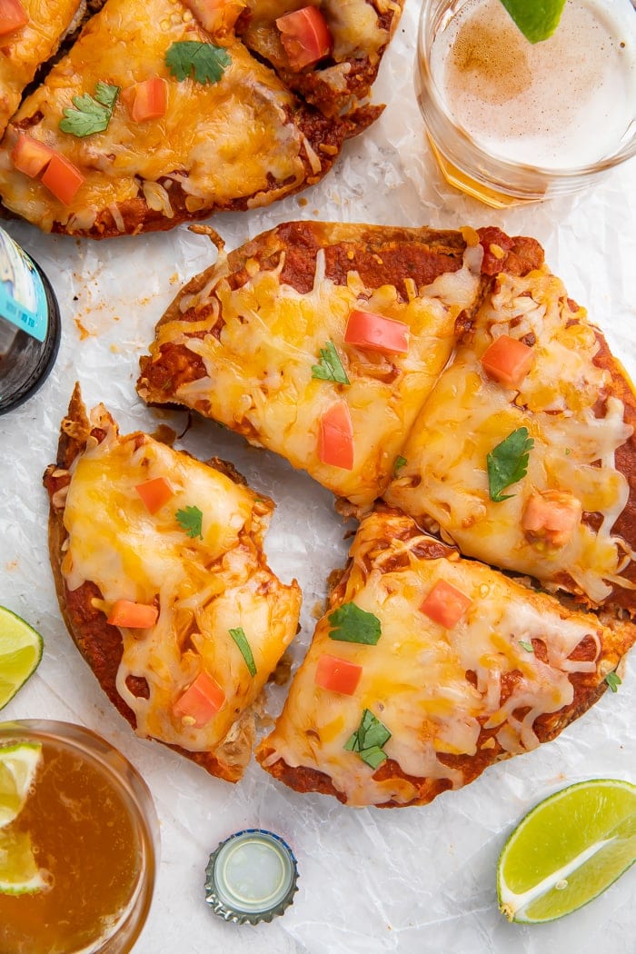 Overhead shot of Taco Bell Mexican pizza recipe topped with diced tomatoes and cilantro, with one slice taken out with a bite in it, surrounded by a lime wedge, glass of beer, and a bottle cap