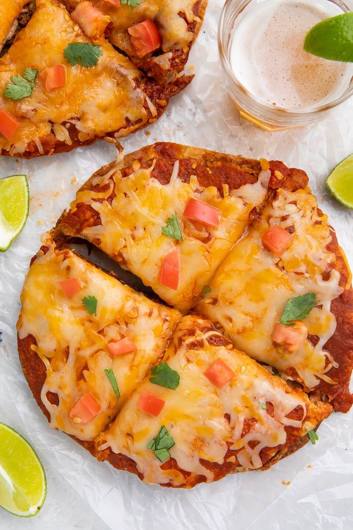 Overhead shot of Taco Bell Mexican pizza recipe topped with diced tomatoes and cilantro cut into four slices, surrounded by a lime wedge, glass of beer, and a bottle cap