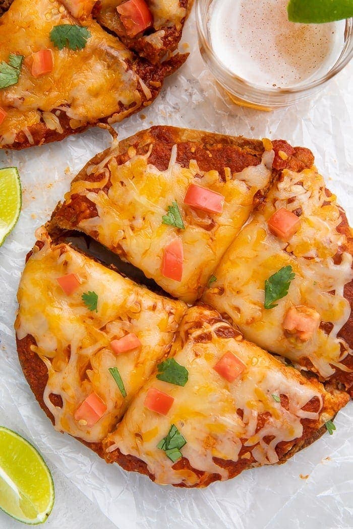 Overhead shot of Taco Bell Mexican pizza recipe topped with diced tomatoes and cilantro, surrounded by a glass of beer and lime wedges