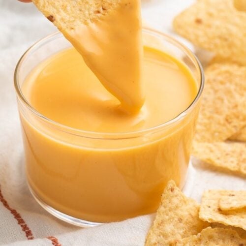 A triangular tortilla chip being dipped into a clear glass of Taco Bell nacho cheese sauce while surrounded by broken tortilla chips