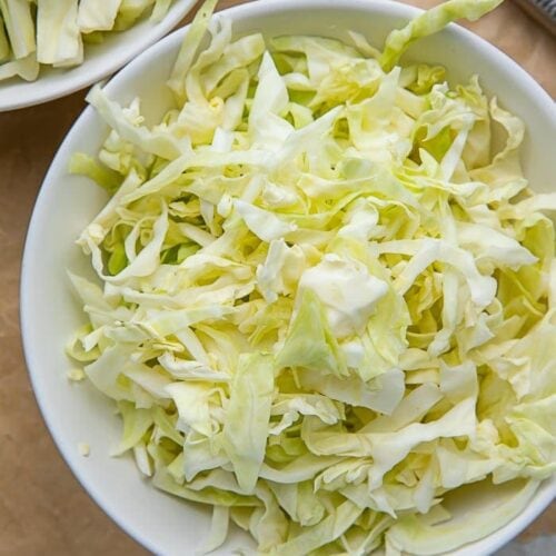 A white bowl full of shredded cabbage on a wooden cutting board next to a knife