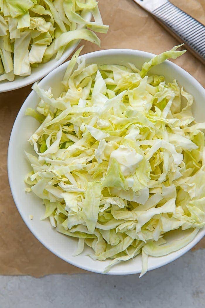 A white bowl full of shredded cabbage on a wooden cutting board next to a knife