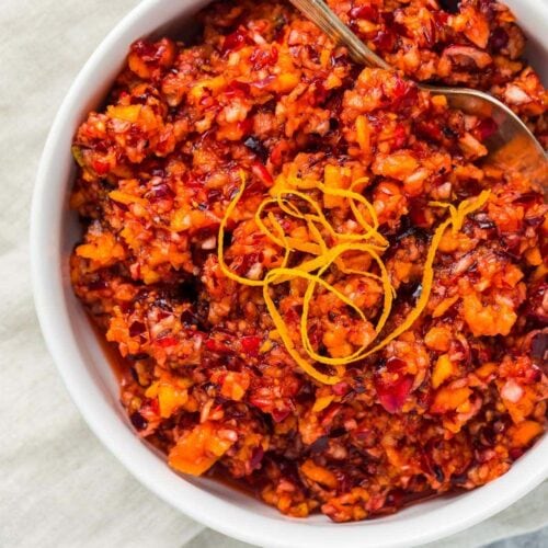 Bright red cranberry relish with orange peel garnish in a white bowl on a napkin and marble countertop