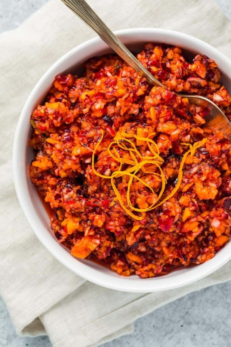 Bright red cranberry relish with orange peel garnish in a white bowl on a napkin and marble countertop