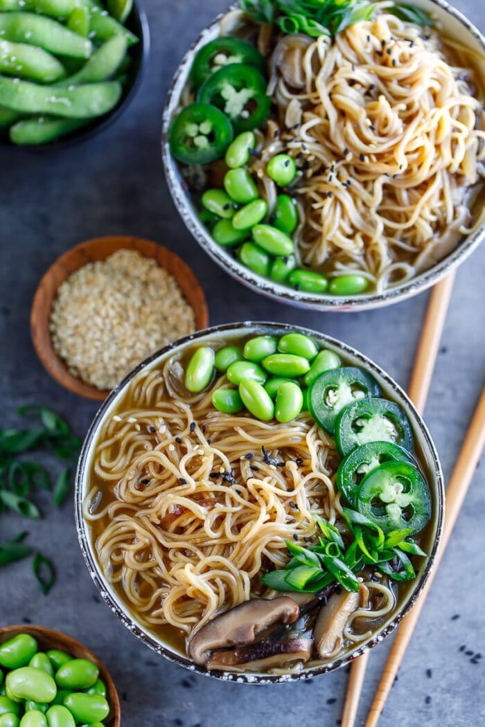 Two black bowls with ramen and veggies sitting on a dark countertop with chopsticks