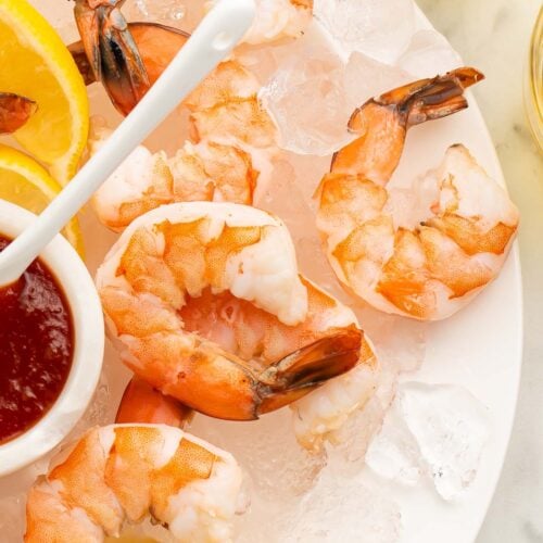 A close-up of poached shrimp arranged on a platter of ice next to a small dipping bowl of shrimp cocktail sauce.