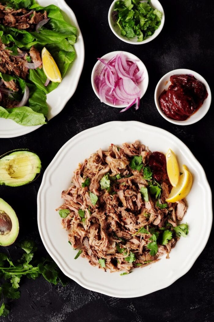 Sous vide Mexican pulled pork in a white bowl next to smaller bowls of garnish for Mexican street tacos