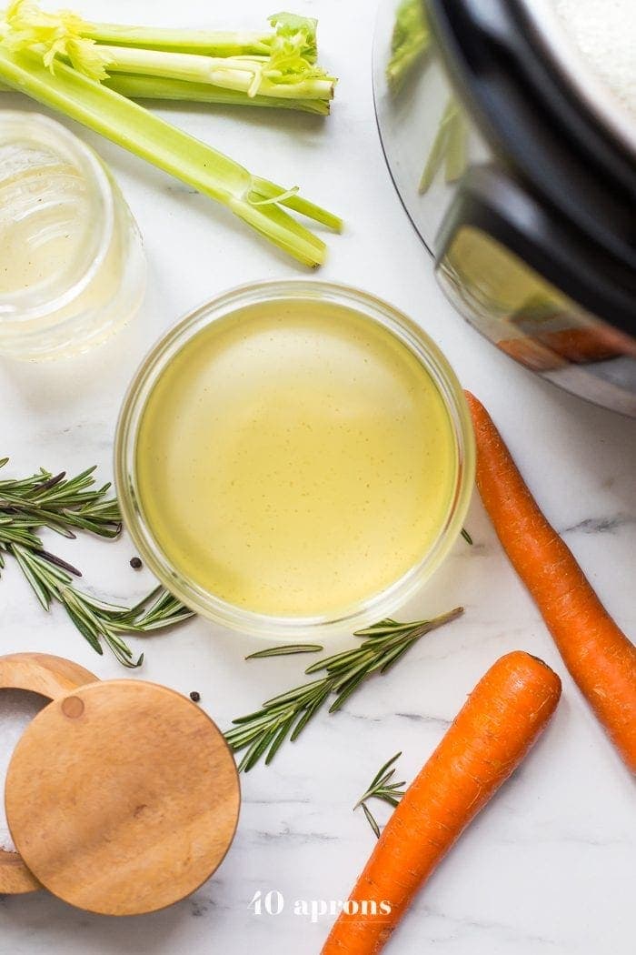 A mason jar of bone broth next to an Instant Pot on a grey countertop. Celery and carrots are scattered around.