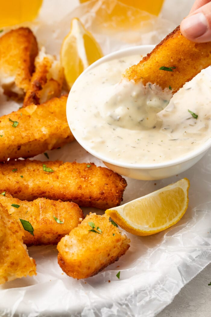 A close up of an air fried fish stick being dipped into a small bowl of dipping sauce 