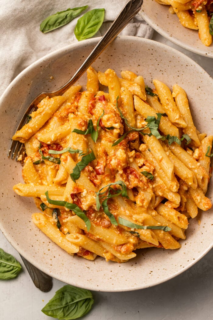 Baked feta pasta in a speckled ceramic bowl, with a spoon, surrounded by fresh basil leaves