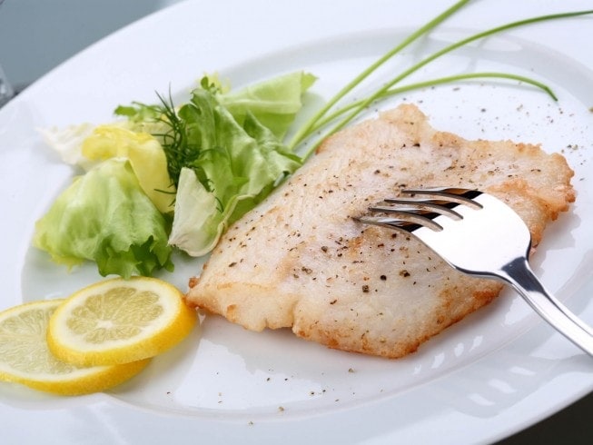 Grilled orange roughy on a white plate with lemon slices and a small salad