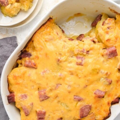 Overhead angle of cheesy ham and potato casserole in a white casserole dish, with one serving scooped onto a white plate