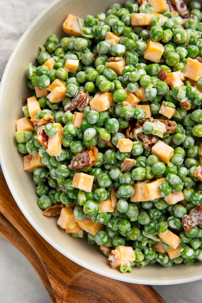 Overhead photo of pea salad with bacon, cheddar cheese, and green onion in a large bowl next to wooden utensils