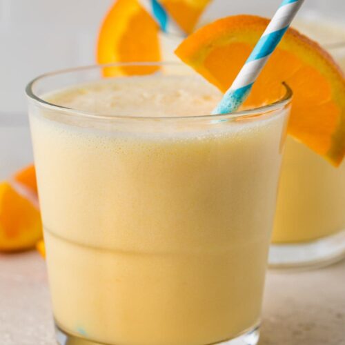 Short glass containing a serving of Orange Julius with an orange slice and a blue and white striped straw