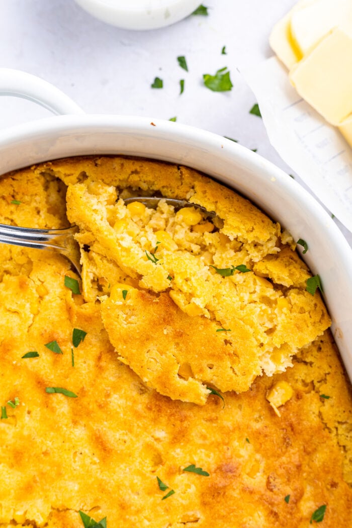 Overhead image on cornbread casserole being scooped out of baking dish with sliced butter on the side.