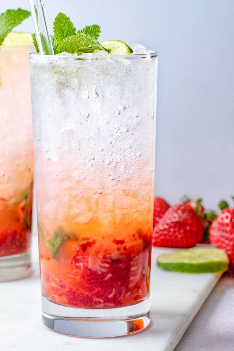 A tall highball glass containing a strawberry mojito, with rum and ice layer on top of muddled strawberries and mint.