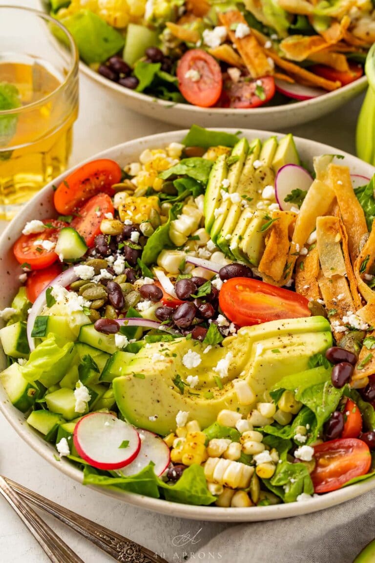 Closeup angle of Mexican salad with slices of avocado, tomatoes, tortilla strips, and onion in a white bowl.